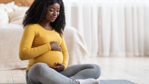 Pregnant woman sitting cross-legged, cradling her belly, wearing a yellow top and gray leggings.