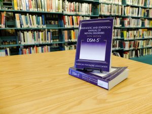 Image of front cover of one DSM-5 manual propped on top of another DSM-5 manual on the table surface. Library bookshelves are in the background.