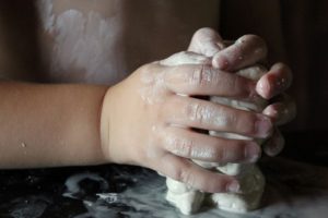 Image of a child’s small hands molding clay on a surface. Clay marks are visible on child’s fingers and nails.