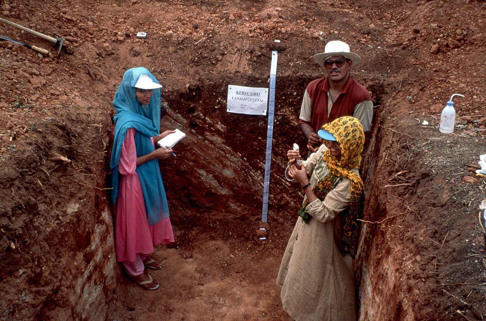 Three people in an excavation trench examining the site with tools and a measurement tape on the wall.