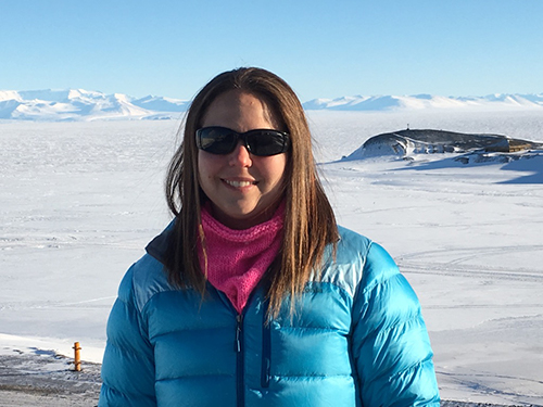 A woman wearing sunglasses, a blue puffy jacket, and a pink neck gaiter stands smiling in a snowy, mountainous landscape under a clear blue sky.