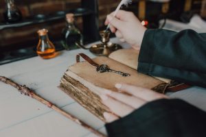A close of a pair of hands dipping a feather quill in an ink pot and preparing to write in a book made of yellowed parchment.