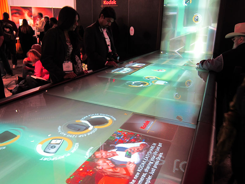 Trade-show visitors interact with Kodak’s “Digital Waterfall,” a long, glowing multi-touch table that streams product images and icons toward a vertical screen. (Image Description available)