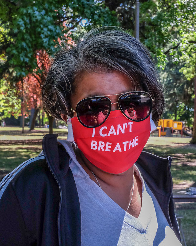 Person outdoors in a park wearing sunglasses and a red face mask that reads “I CAN’T BREATHE.”